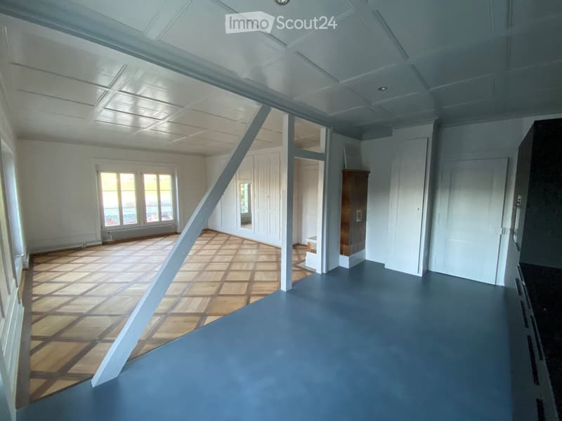 Empty room with wooden flooring and white walls, featuring large windows and a visible doorway leading to another room.
