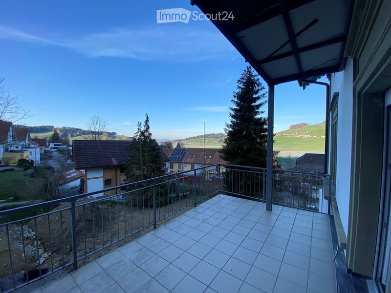 Balcony with tiled floor and metal railing, overlooking a rural landscape with houses, trees, and open fields.