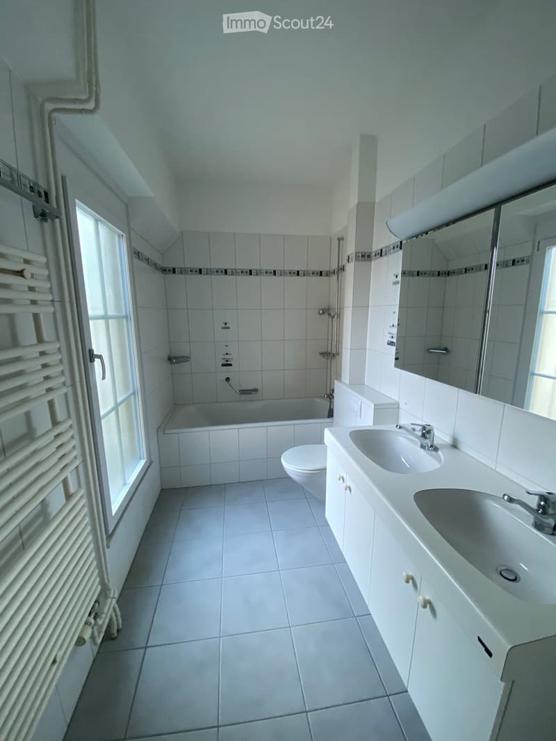 Modern bathroom with white tiles, a double sink vanity, toilet, bathtub, and a large mirror above the sink. A radiator is mounted on the wall near the window.
