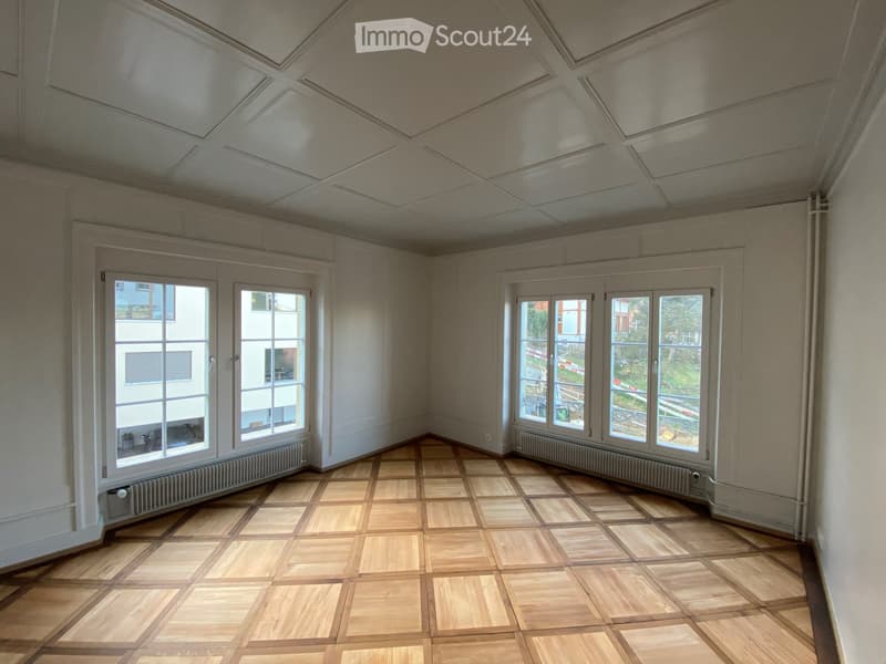 Empty room with wooden parquet flooring, white walls, large windows, and radiators. No furniture or equipment present.