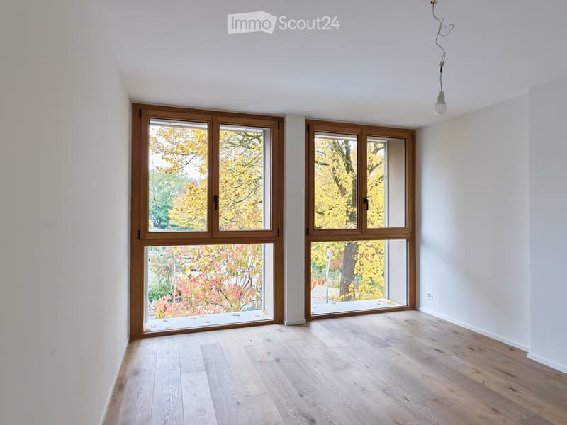 Empty room with light wooden flooring, white walls, and large windows with wooden frames offering views of autumn foliage.