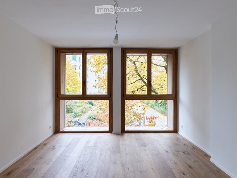 Empty room with light wooden flooring and large windows offering views of trees and a pathway.