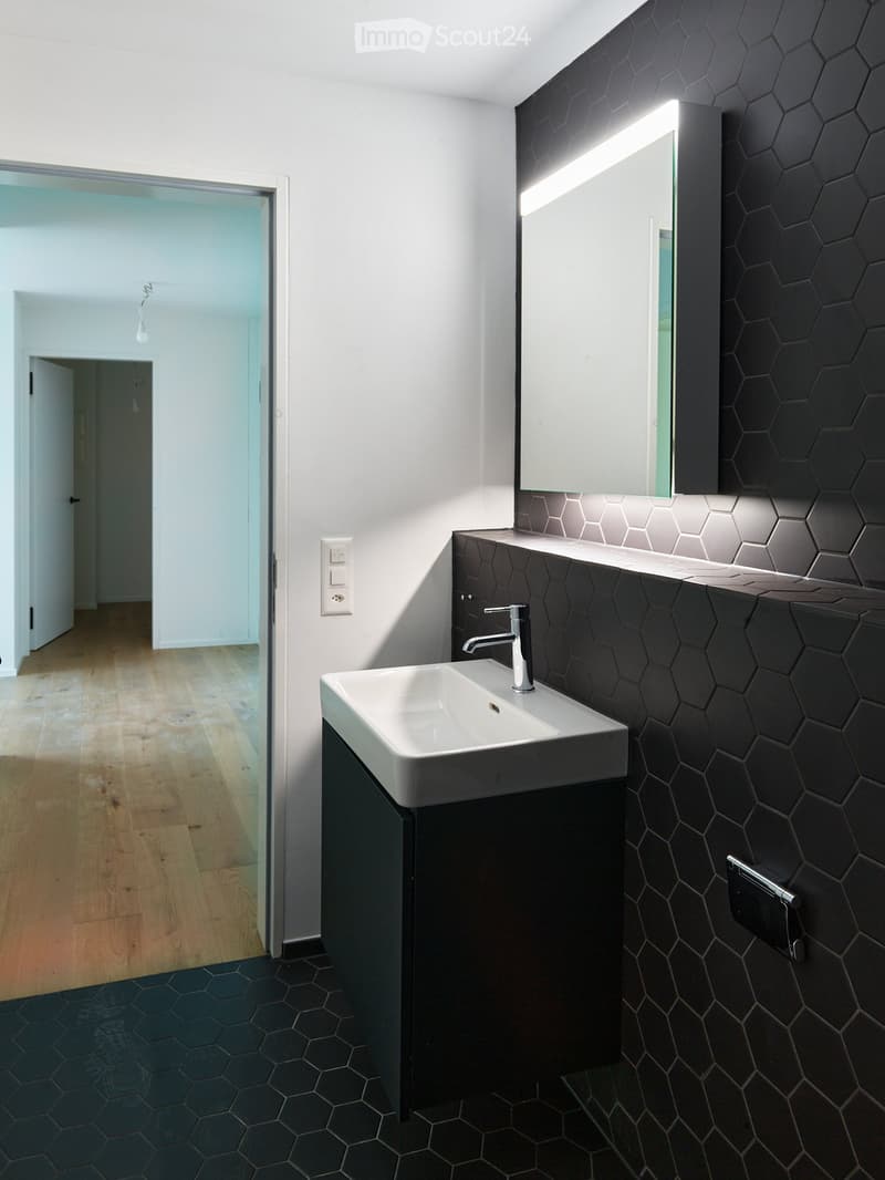 Modern bathroom with hexagonal black tiles on walls and floor, white sink with chrome faucet, large mirror with built-in lighting, and dark wooden cabinet.