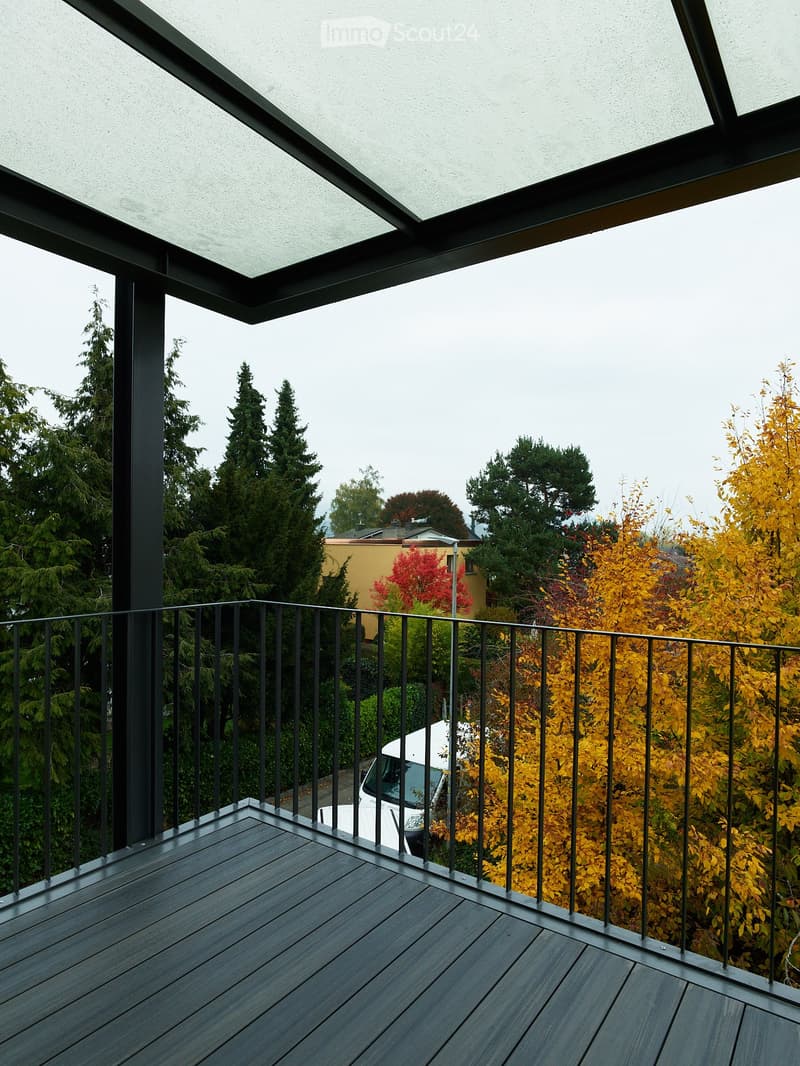 Modern balcony with dark wooden decking, black metal railing, and a retractable roof, overlooking a lush, green landscape with trees and a parked car below.