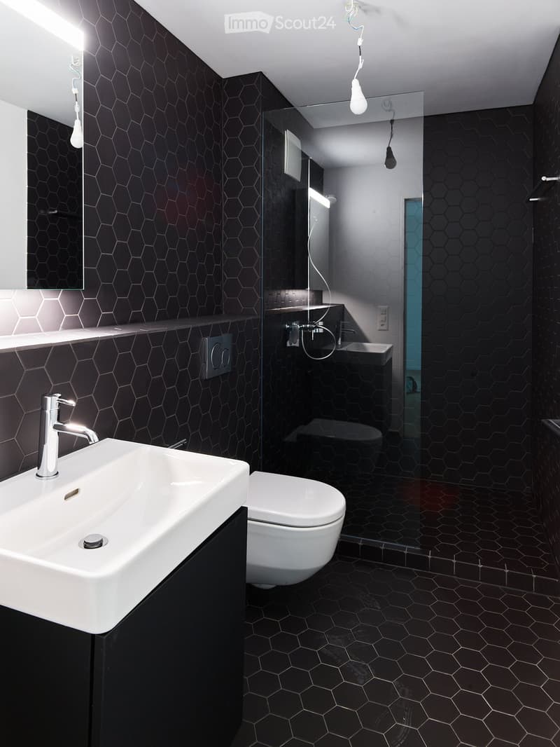 Modern bathroom with hexagonal black tiles, white sink, chrome faucet, toilet, and glass-enclosed shower area.