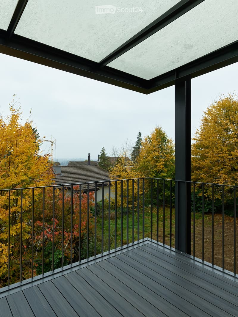 Modern balcony with dark wooden decking, black metal railing, and a retractable roof, overlooking a landscape with autumn-colored trees and a distant house.
