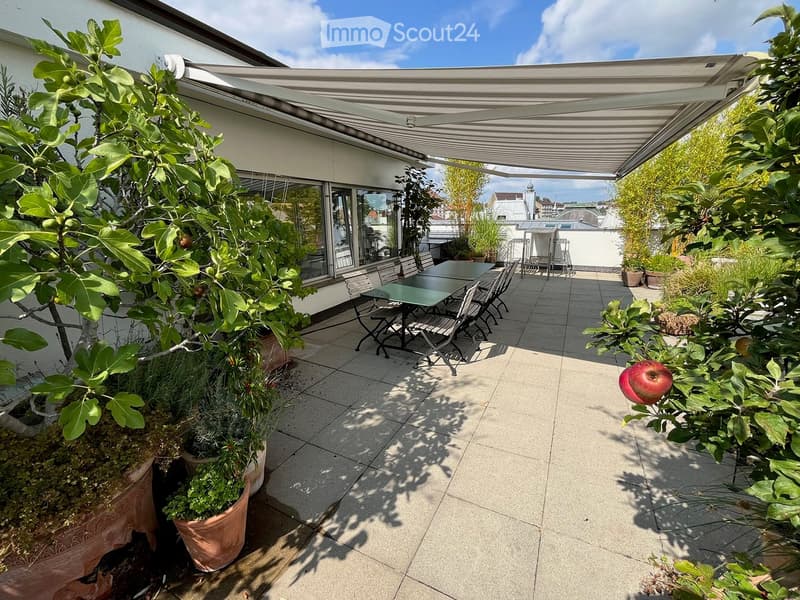 A spacious, covered balcony with a tiled floor, a dining table and chairs, surrounded by potted plants and greenery, with a view of the city.