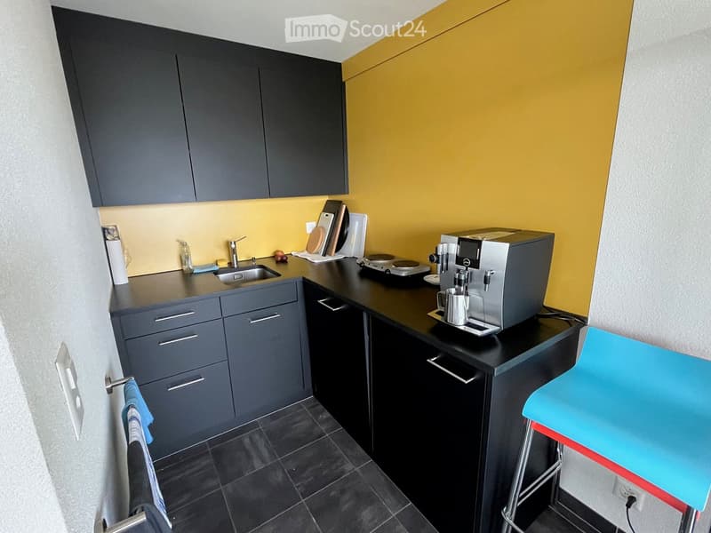 Modern kitchen with dark grey cabinets, black countertops, a sink, a coffee machine, and a stool. The walls are painted in a bright yellow color, and the floor is tiled in dark grey.