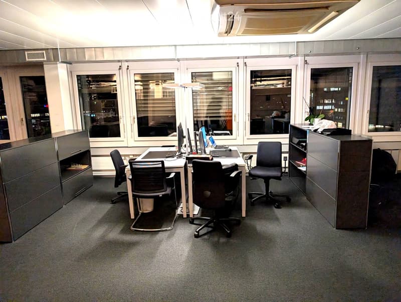 Office space with desks, chairs, laptops, and computer monitors, large windows with blinds, and a cabinet.