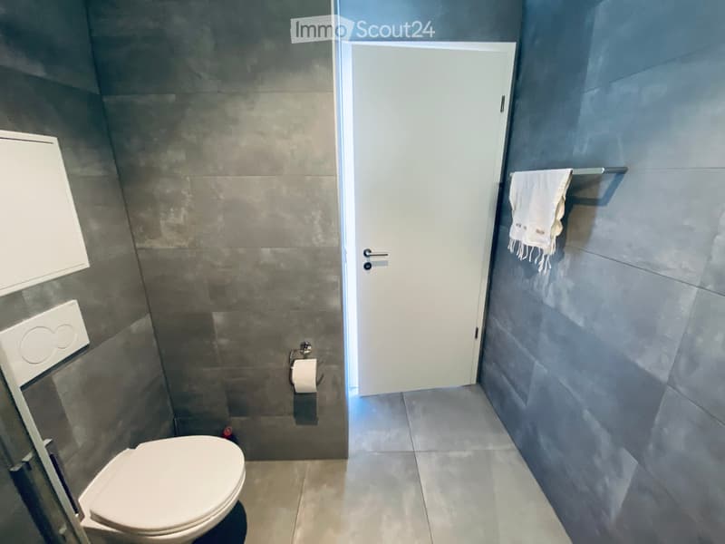 Modern bathroom with grey tiled walls, white toilet, towel on a rack, and a cabinet.