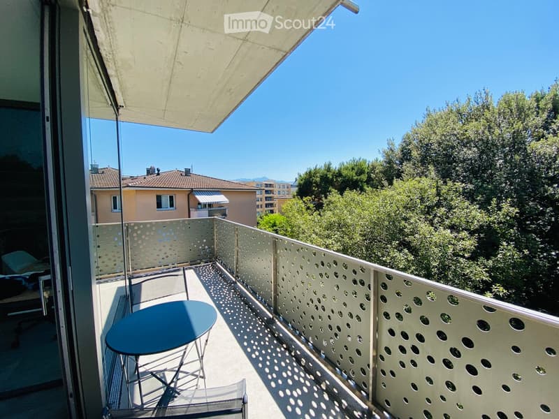 Balcony with a table and chair, featuring a perforated metal railing and a large umbrella providing shade. The view includes trees and neighboring buildings.