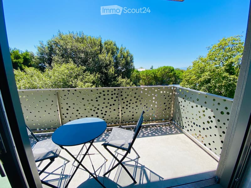Balcony with a round blue table and two folding chairs, surrounded by a perforated metal railing and trees in the background.