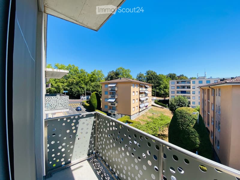 Balcony with metal railing featuring circular cutouts, overlooking a residential area with buildings, trees, and a clear blue sky.