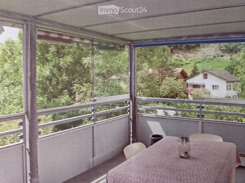 Balcony with a table and chairs, overlooking a green, hilly landscape with houses and trees.
