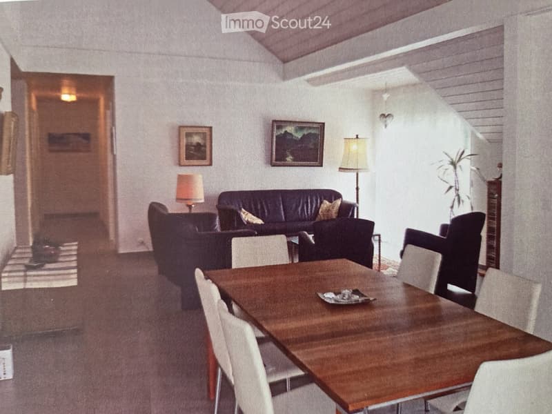 A cozy living room with a dark leather sofa, a wooden dining table with chairs, and two lamps. The room features white walls, a slanted ceiling, and framed artwork.