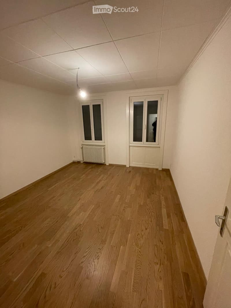 Empty room with wooden flooring, white walls, and two windows with white frames. A radiator is visible under one window, and a door is on the right side.