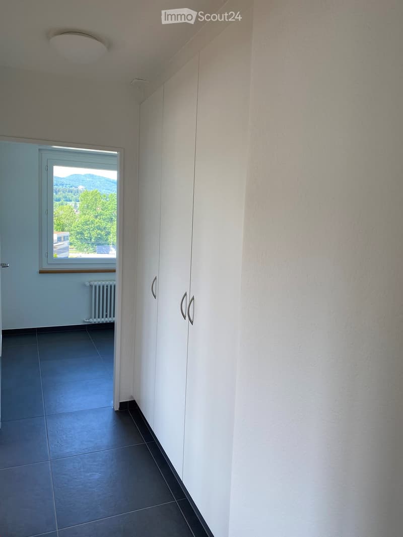 Empty hallway with white walls, dark tiled floor, and a window showing a view of greenery and mountains.