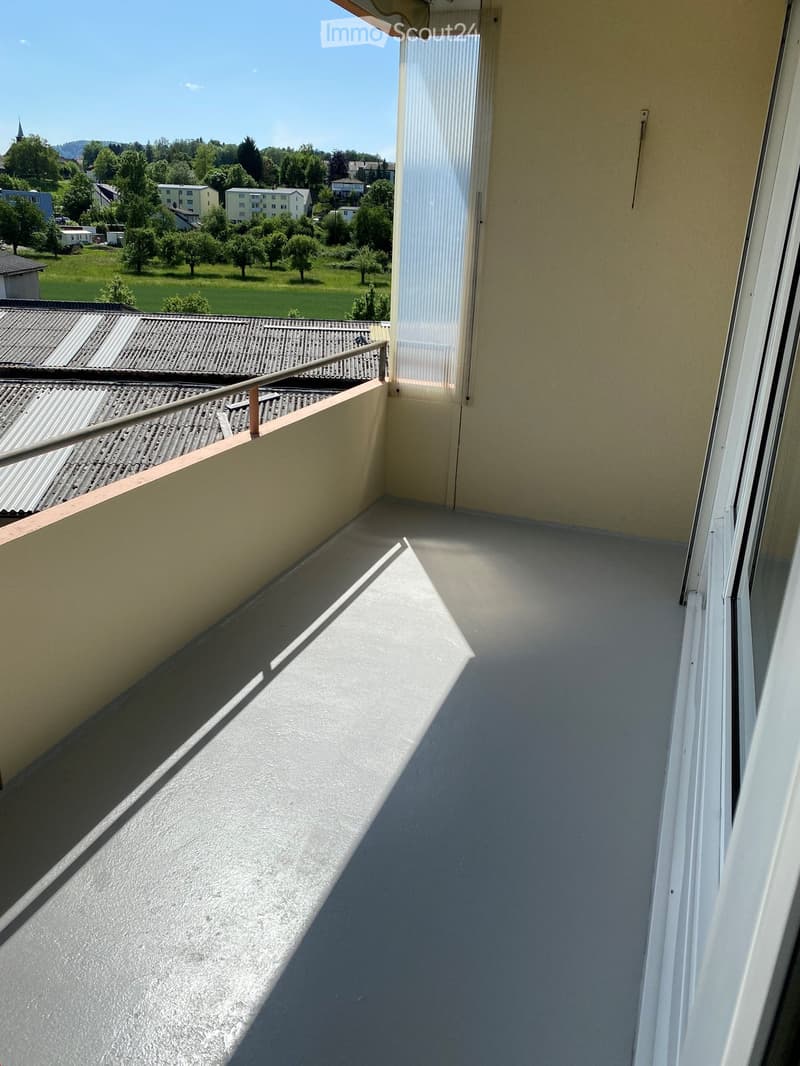 Balcony with a metal railing, overlooking a green landscape with trees and houses.