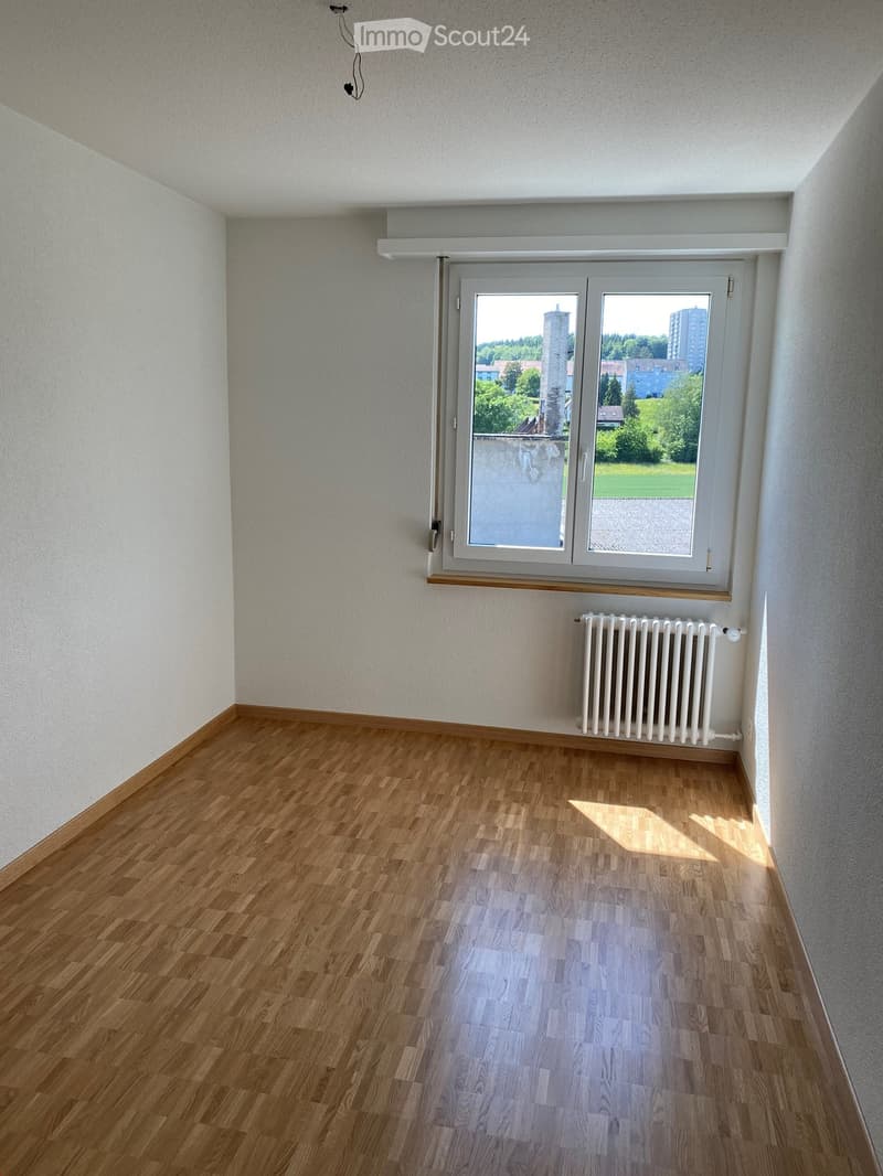 Empty room with wooden flooring, white walls, and a large window offering a view of greenery and buildings. A radiator is visible below the window.