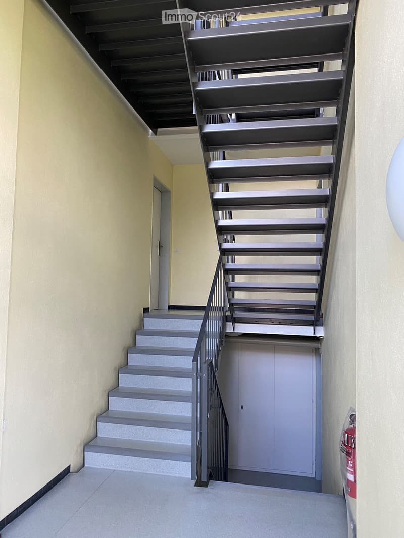 Modern interior staircase with gray steps and metal railing, leading to an upper level. Beige walls and tiled floor.