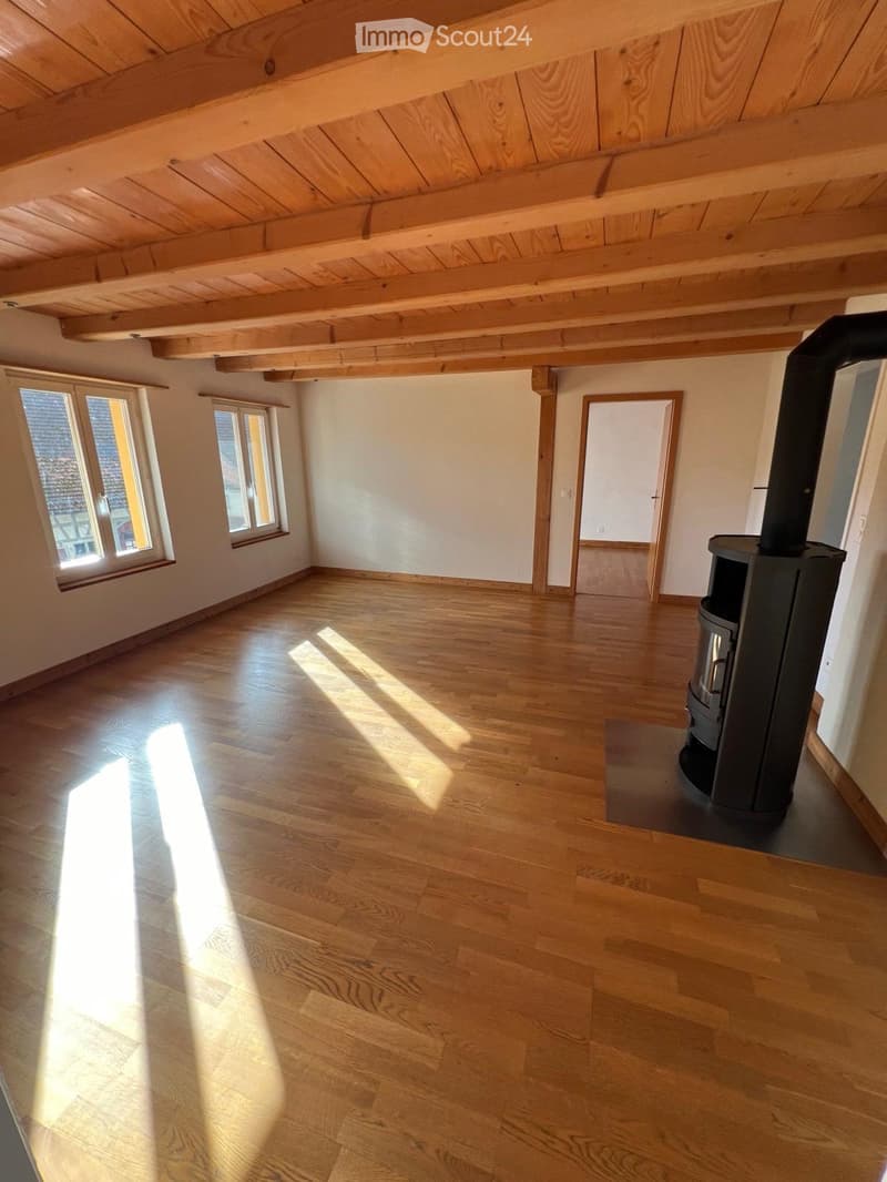 Empty room with wooden floor, exposed wooden ceiling beams, large windows, and a black wood-burning stove in the corner.