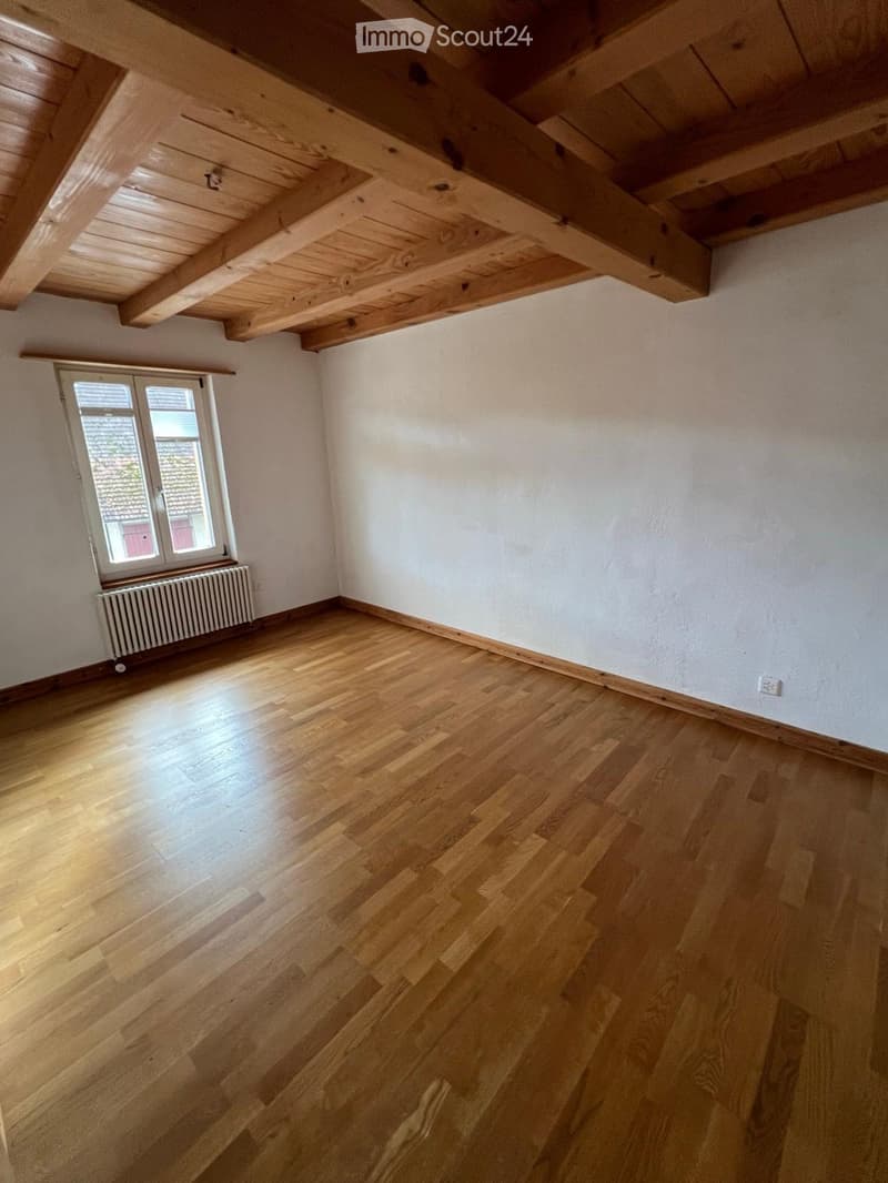 Empty room with wooden flooring, white walls, exposed wooden ceiling beams, and a single window with a radiator below it.