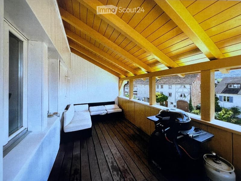Wooden balcony with a covered roof, featuring a sofa, grill, and a small table with utensils.