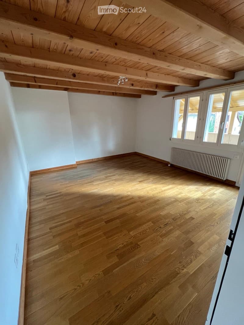 Empty room with wooden floor, exposed wooden ceiling beams, light blue walls, and a radiator. Windows provide natural light.