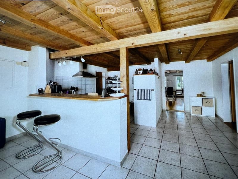 Open-plan kitchen and living area with exposed wooden beams, white walls, tiled floor, and modern appliances.