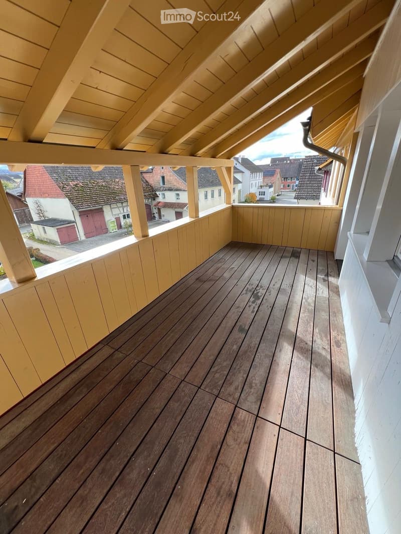 Wooden balcony with slatted floor, wooden railings, and visible neighborhood houses in the background.