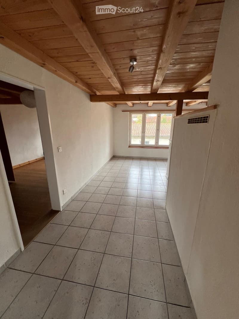 Empty room with exposed wooden beams on the ceiling, tiled floor, and large windows letting in natural light.