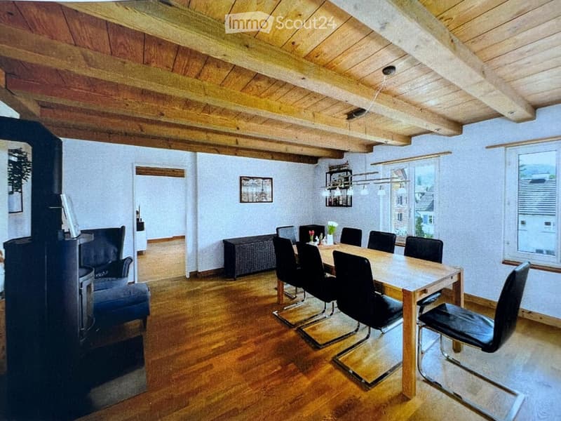 A cozy living and dining area with exposed wooden beams, a fireplace, a sofa, a dining table with chairs, and a chandelier.