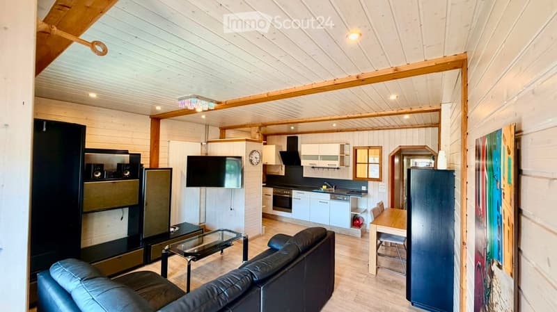 Open-plan living and dining area with a leather sofa, coffee table, TV, and a small kitchen area with cabinets, oven, and refrigerator. Wooden ceiling beams and warm lighting.