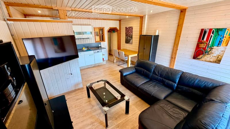 A modern, open-concept living and dining area with wooden walls and ceiling beams. Features a black leather sofa, glass coffee table, flat-screen TV, light blue kitchen cabinets, dining table with chairs, and a refrigerator.
