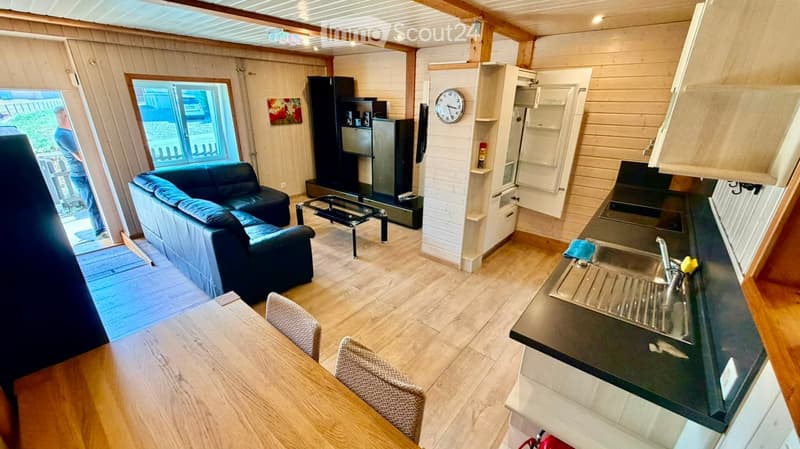 Open-plan living and dining area with a sofa, coffee table, dining table with chairs, kitchen counter with sink, and a small fridge. Wooden walls and ceiling, large windows, and a clock on the wall.