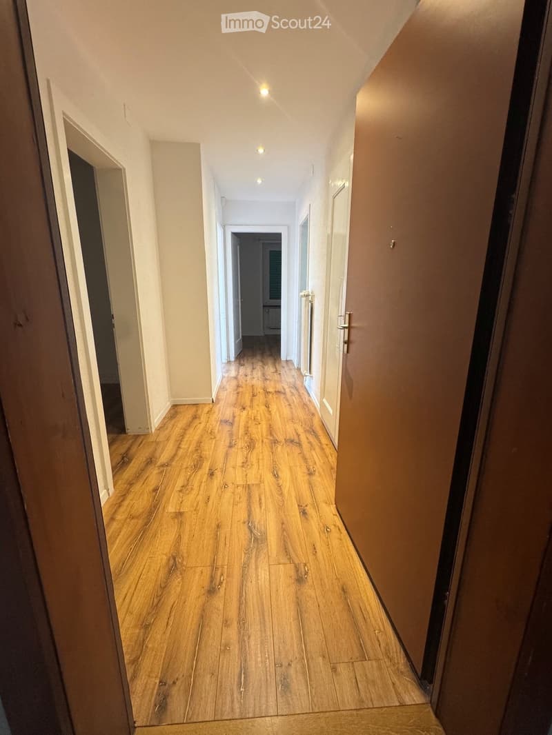 A long, narrow hallway with wooden flooring and white walls, featuring several doors on both sides and recessed ceiling lighting.
