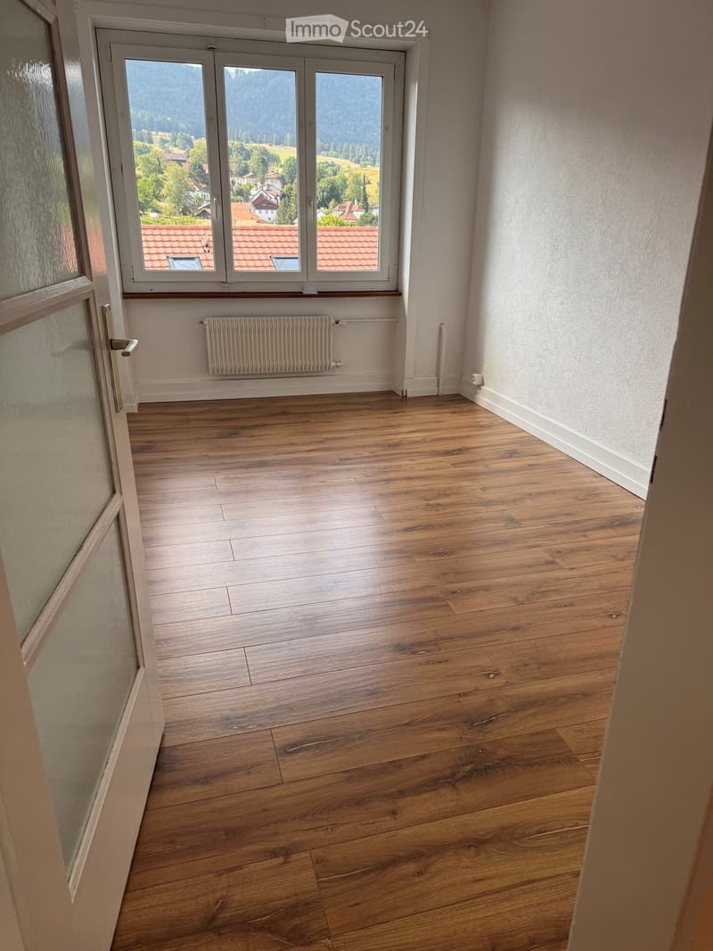 Empty room with wooden flooring, white walls, a radiator, and large windows offering a scenic view of a village and mountains.