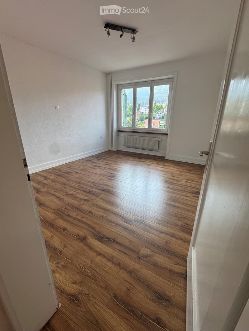 Empty room with wooden flooring, white walls, large window with view, radiator, and ceiling spotlights.
