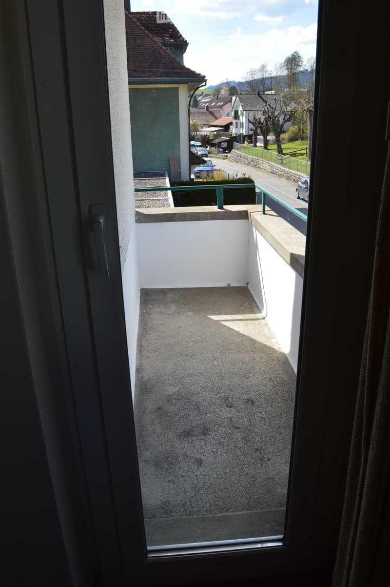 Balcony with concrete floor, metal railing, overlooking a street with houses, trees, and parked cars.