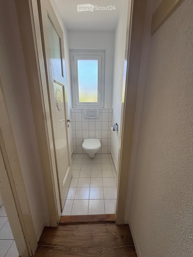 Small bathroom with white tiled walls and floor, toilet, window, and a partially open door leading to a hallway.