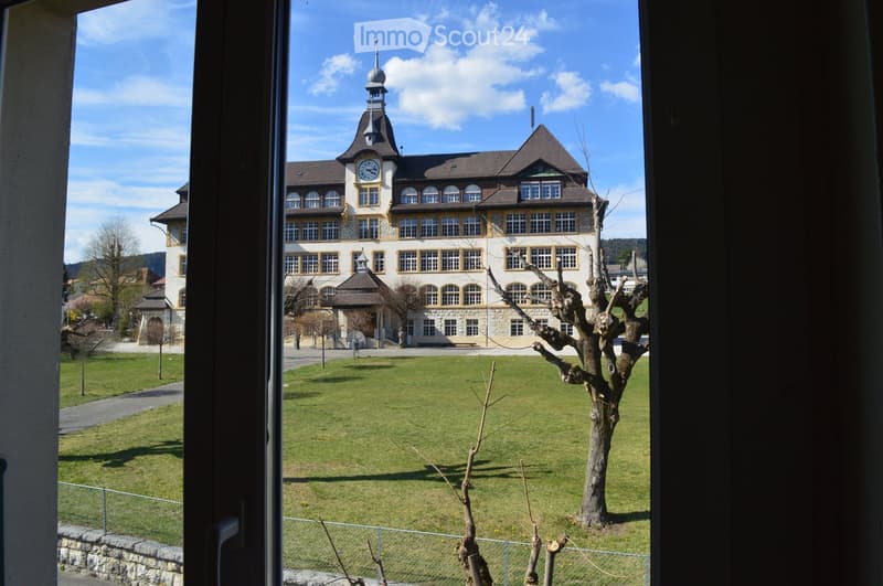 View from a window showing a large, historic building with a clock tower, surrounded by a well-maintained lawn and trees.