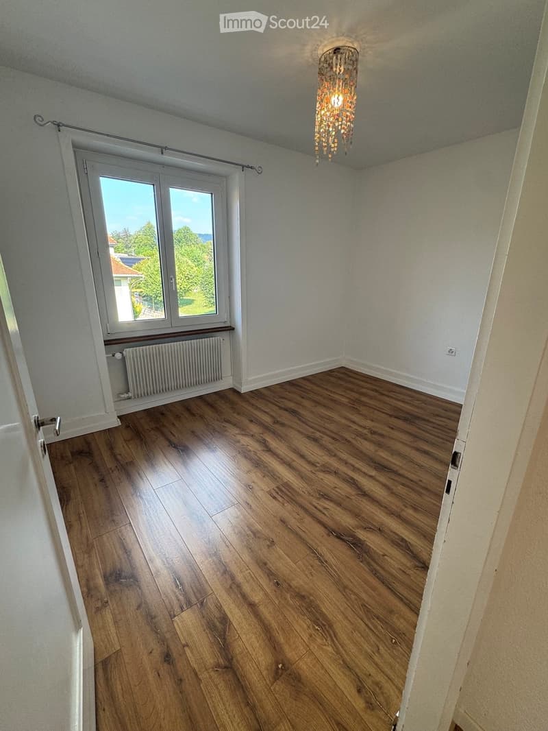 Empty room with wooden flooring, white walls, large window with view, radiator, and chandelier.