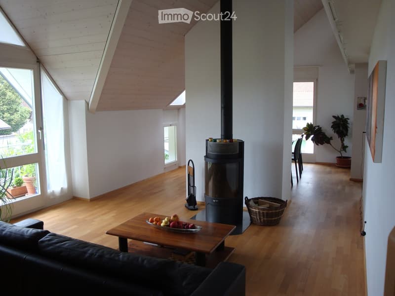 Bright living room with wooden floors, white walls, and large windows. Features a black leather sofa, wooden coffee table with fruit bowl, modern wood-burning stove, and a wicker basket on the floor.