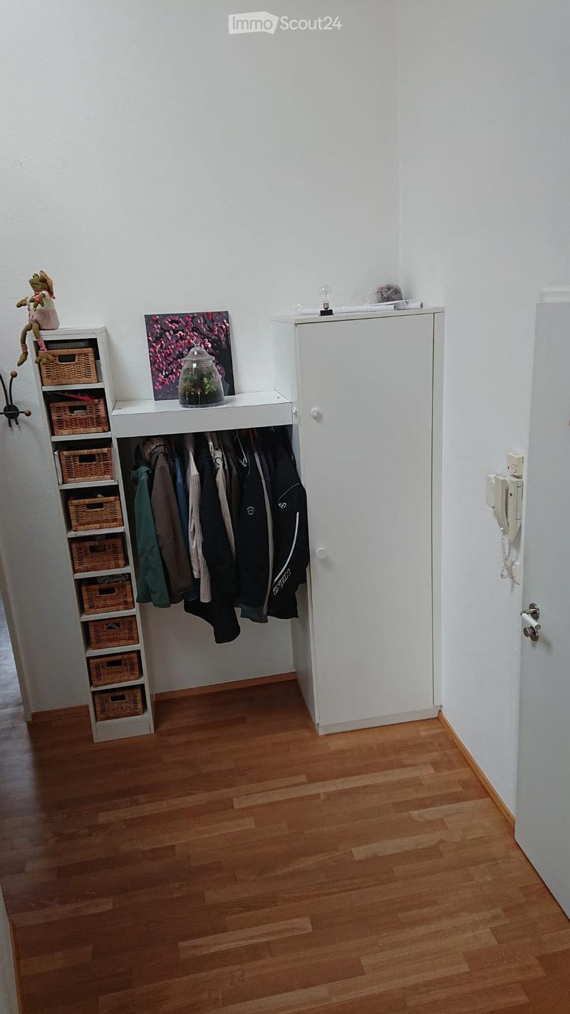 A small hallway with wooden flooring, white walls, a built-in closet with hanging clothes, a shelf with wicker baskets, and a small decorative display including a framed picture and a glass jar with a plant inside.