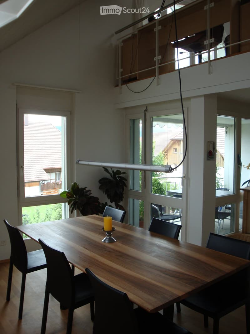 Bright, open dining area with large windows and a wooden table surrounded by black chairs. A yellow candle sits in the center of the table. The room features modern lighting and visible greenery in the background.