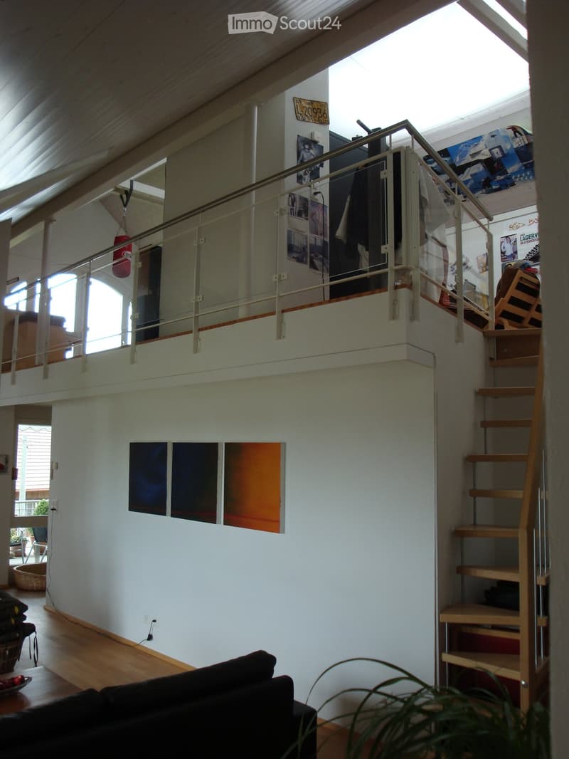 Open-plan living space with wooden flooring, a staircase leading to an upper level, a sofa, a small table, a plant, and shelves with various items.