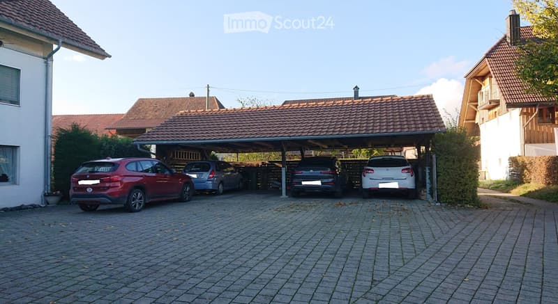 Driveway with four parked cars under a carport attached to a house with a tiled roof.