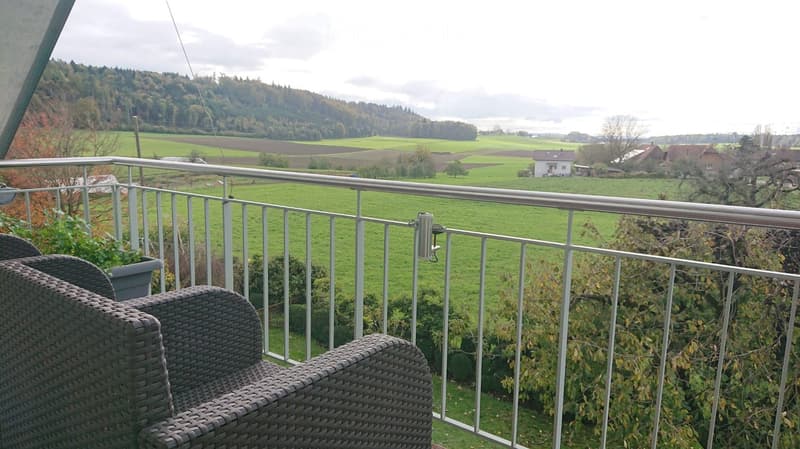 Balcony with metal railing, overlooking a green field and forested hills, with wicker chairs and a small table.