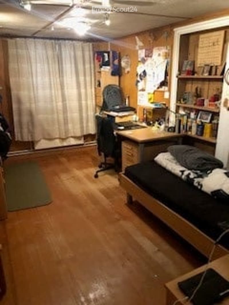 A small, cozy room with wooden flooring, a desk with a chair, a bed, a bookshelf, and a laptop on a small table. The room has a curtain covering a window and a rug on the floor.