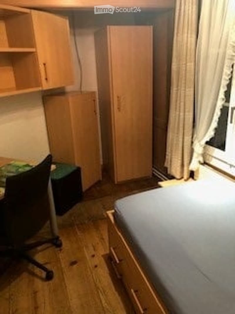 A small, furnished bedroom with a single bed, a chair, a desk, a wardrobe, and a cabinet. The room has wooden flooring and white walls.